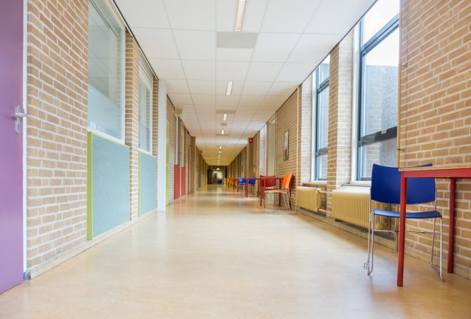 Long empty corridor with furniture in school building. Colorful hall with brick walls and windows. To the left there are many classrooms with doors. Children and teenagers have to walk through this corridor to attend lessons in class.